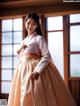 A woman in a white and orange hanbok standing in front of a window.