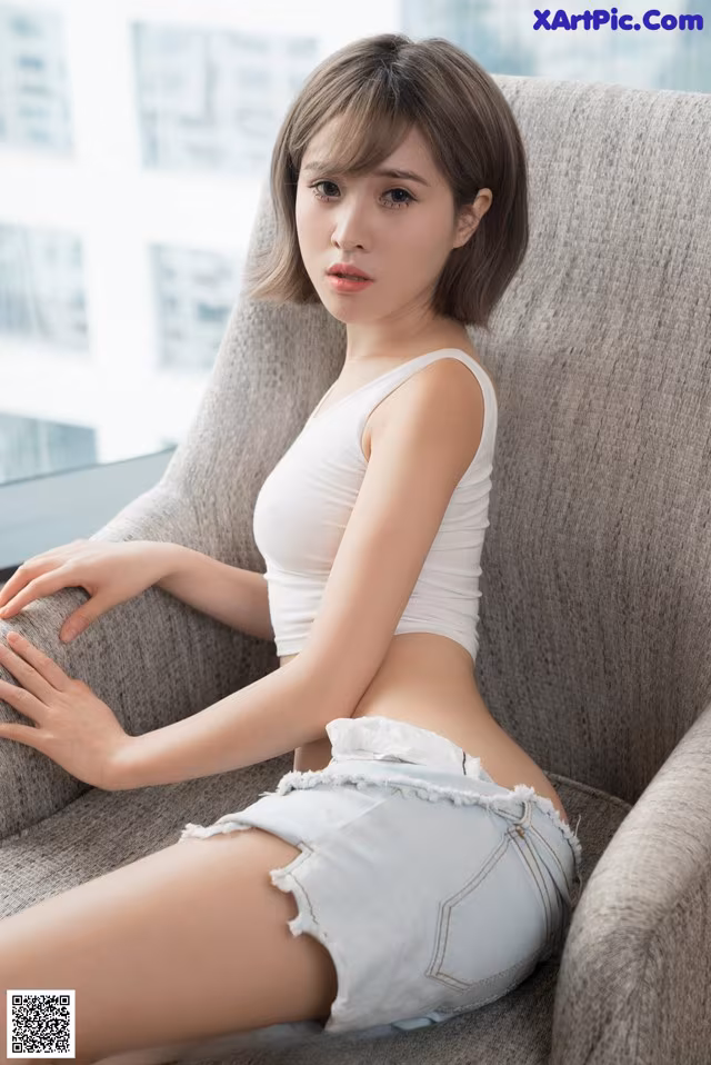 A woman sitting on a chair in a white top and denim shorts.